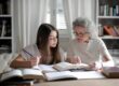 A grandmother helps her teenage granddaughter with homework, surrounded by books indoors.